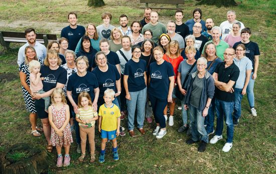 Das Team von Gut Wilhelmsdorf. Sommerlich gekleidet in dunkel blauen Gut Wilhelmsdorf T-Shirts und in Alltagskleidung. Kinder stehen ganz vorne oder werden getragen.