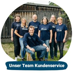 KI generiert: Das Bild zeigt eine Gruppe von sieben Personen in blauen T-Shirts, die vor einer alten Holzwand im Freien posieren. Der Mann in der Mitte sitzt auf einem Holzstamm, während die anderen um ihn herum stehen.