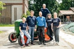 KI generiert: Eine Gruppe von sechs Personen posiert lächelnd bei sonnigem Wetter vor einem landwirtschaftlichen Gebäude neben einem grünen Traktor. Sie tragen überwiegend dunkelblaue Kleidung und scheinen Teil eines Teams oder einer Gemeinschaft zu sein.