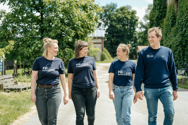 KI generiert: Vier Personen, die im Freien gemeinsam nebeneinander laufen und dabei lächeln. Sie tragen ähnliche blaue T-Shirts mit dem Aufdruck "Gut Wilhelmsdorf".