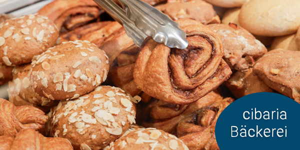 KI generiert: Das Bild zeigt eine Auswahl an frischem Gebäck, darunter mit Mandeln bestreute Brötchen und eine Zange, die eine Zimtschnecke hält. Im Hintergrund sind weitere Backwaren zu sehen.