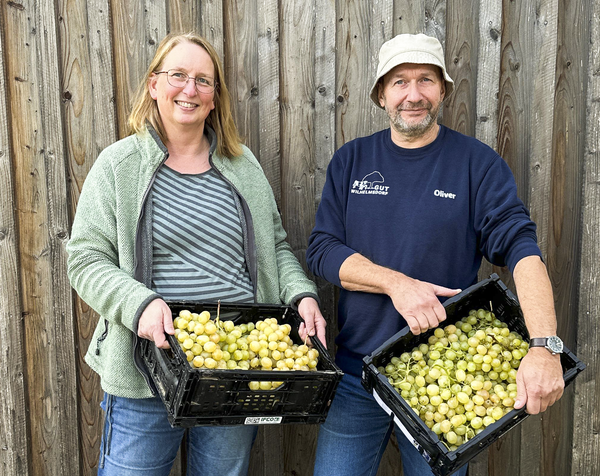 Tanja und Oli halten lächelnd vor einer Holzwand Kisten mit selbst geernteten Trauben von der Plantage von Don Luigi auf Sizilien.