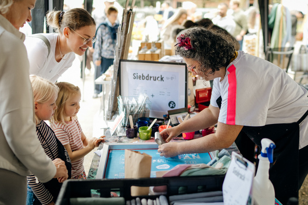 KI generiert: Person am Marktstand zeigt Kindern Siebdruck. Text: "Siebdruck to-go".