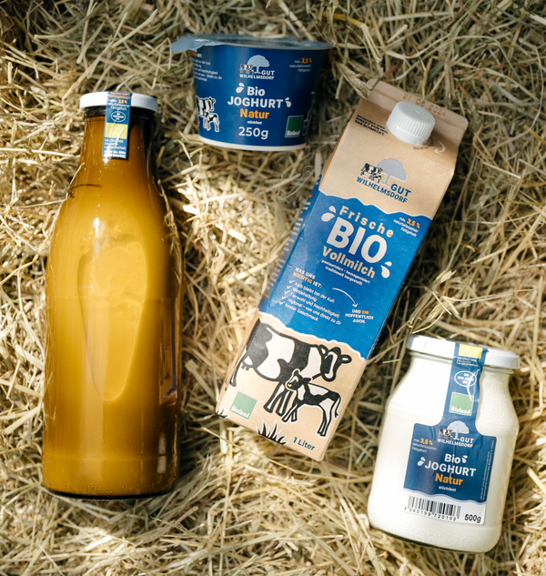Die Produkte unserer regionalen Hofmolkerei, Milch in der Flasche und im Karton, Joghurt im Glas und im Becher, auf einem Strohbett arrangiert.
