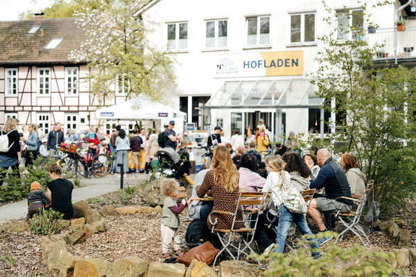 KI generiert: Menschen sitzen und stehen vor einem Hofladen. Text: "HOFLADEN".
