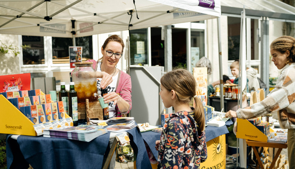 KI generiert: Eine Frau serviert Getränke an einem Marktstand mit Sonnentor-Produkten.