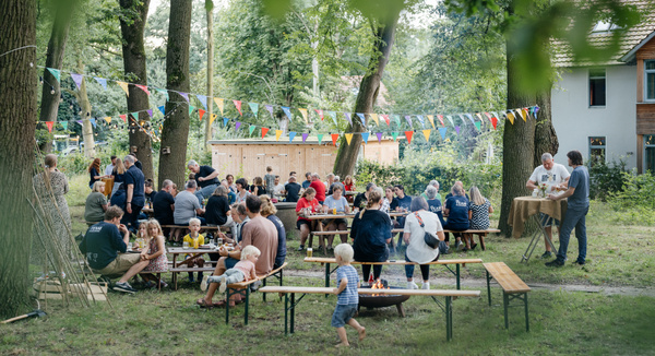 KI generiert: Das Bild zeigt ein geselliges Treffen im Freien, bei dem Menschen auf Holz- und Bierbänken unter bunt beflaggten Girlanden sitzen. Es wirkt wie ein entspanntes Sommerfest in einem bewaldeten Garten oder Park.