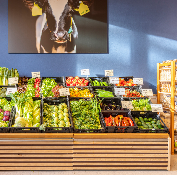 KI generiert: Marktstand mit frischem Gemüse in Kisten. Im Hintergrund ein Bild einer Kuh.