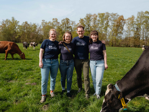 KI generiert: Vier Personen stehen auf einer Weide mit Kühen. Texthinweis: „WILHELM GUT WILLENBROCKS“.