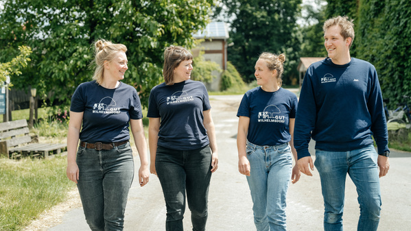 KI generiert: Vier Personen, die im Freien gemeinsam nebeneinander laufen und dabei lächeln. Sie tragen ähnliche blaue T-Shirts mit dem Aufdruck "Gut Wilhelmsdorf".