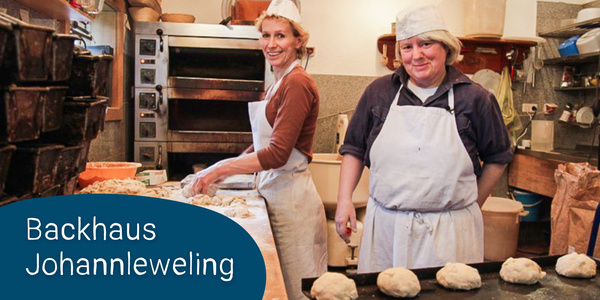 KI generiert: Das Bild zeigt zwei Bäcker in einer Bäckerei bei der Arbeit, umgeben von Teig und Öfen. Im Vordergrund ist ein Schild mit der Aufschrift "Backhaus Johannleweling" zu sehen.