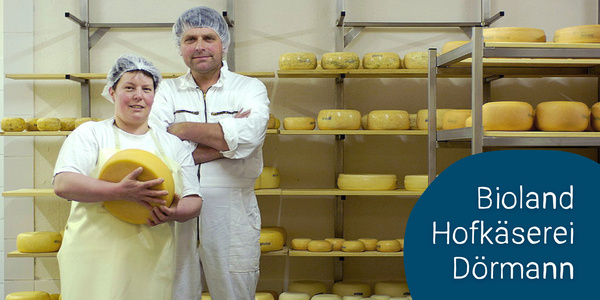 KI generiert: Das Bild zeigt zwei Personen in einer Käserei, die von mehreren Käselaiben umgeben sind. Sie tragen Schutzkleidung und Haarnetze und befinden sich in der Bioland Hofkäserei Dörmann.