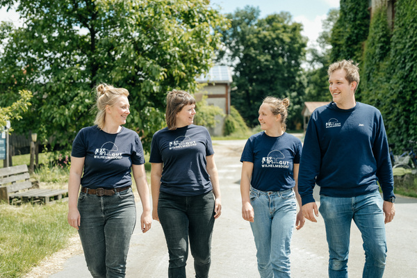 KI generiert: Vier Personen laufen draußen nebeneinander und lächeln. Text auf T-Shirts: "Hofgut Wilhelmsdorf".