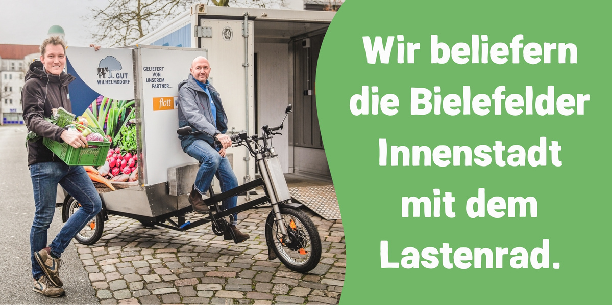 Zwei Personen, Jan und Olli von Gut Wilhelmsdorf, mit einem Lastenrad, das für die Lieferung von Waren in der Bielefelder Innenstadt genutzt wird. Auf dem Lastenrad ist Obst und Gemüse zu sehen und daneben steht ein Slogan, der ihre Lieferdienste bewirbt.