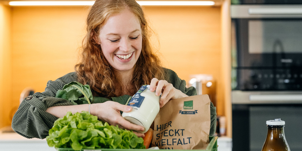 KI generiert: Eine Frau packt frische Lebensmittel aus. Text: "HIER STECKT VIELFALT".