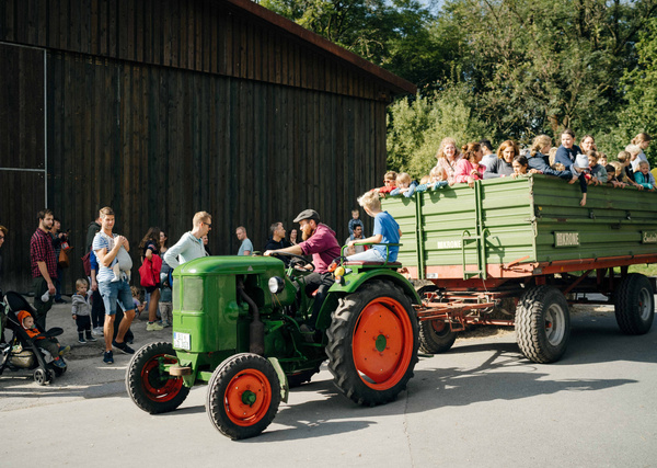 KI generiert: Ein grüner Traktor zieht einen Anhänger voller Menschen vorbei an einem Gebäude.