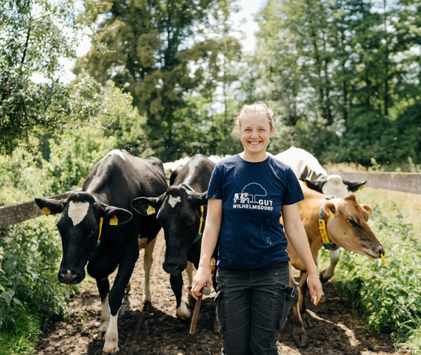 KI generiert: Frau mit Kühen auf einem Feldweg. Text auf Shirt: "GUT WILHELMSDORF".
