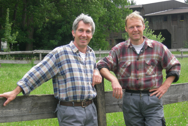 KI generiert: Zwei Männer in karierten Hemden lehnen an einem Holzzaun auf einer Wiese.