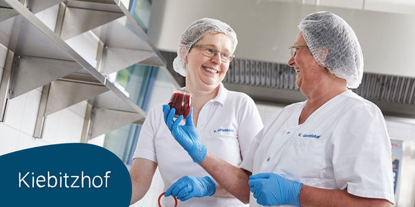 KI generiert: Das Bild zeigt zwei lächelnde Personen in einer Küche, die Schutzkleidung und Handschuhe tragen und ein Glas mit einer roten Flüssigkeit halten. Die Szene vermittelt eine freundliche und kooperative Arbeitsatmosphäre.