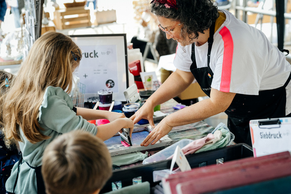 KI generiert: Person hilft Kindern bei einem Siebdruck-Projekt. Text: "Siebdruck to-go".