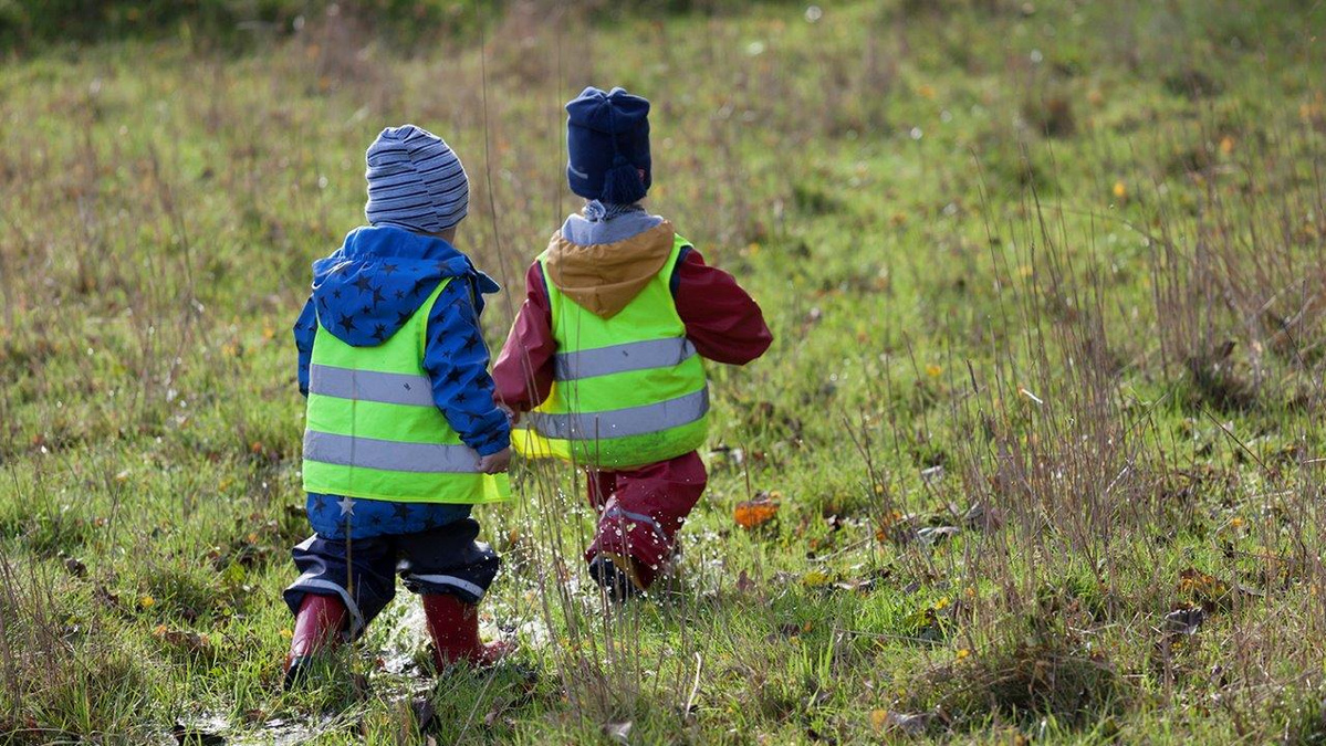 KI generiert: Zwei Kinder in Regenkleidung und Warnwesten gehen über eine Wiese.