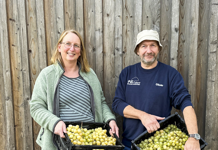 Tanja und Oli halten lächelnd vor einer Holzwand Kisten mit selbst geernteten Trauben von der Plantage von Don Luigi auf Sizilien.
