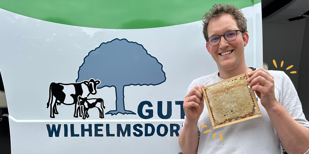 KI generiert: Eine Person hält Honigwabe vor einem Schild mit "Gut Wilhelmsdorf" und Kuh-Baum-Logo.