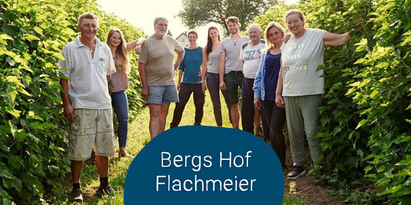 KI generiert: Eine Gruppe von neun Menschen steht lächelnd zwischen hohen Pflanzenreihen auf einem Hof. Im Vordergrund ist der Text "Bergs Hof Flachmeier" zu sehen.