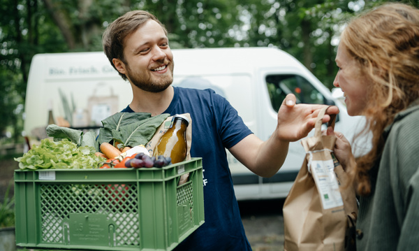 Ein Gut Wilhelmsdorf Auslieferfahrer liefert einer Kundin ihre Biokiste und Wilhelmsdorfer Kartoffeln direkt an die Haustür.