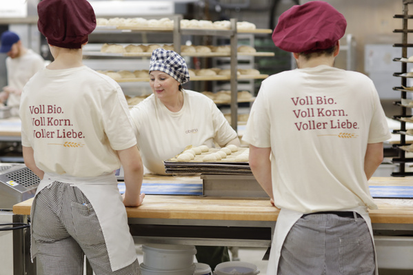 KI generiert: Bäcker formen Teig in einer Bäckerei. Text: "Voll Bio. Voll Korn. Voller Liebe."